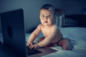 A baby uses a computer while sitting on a bed at night.